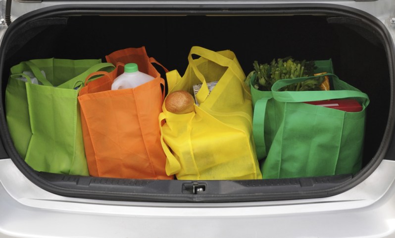 Four colorful eco-friendly shopping bags filled mostly with groceries in the opened trunk of a car.