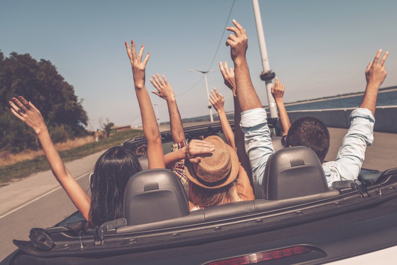 Enjoying road trip. Rear view of young happy people enjoying road trip in their convertible and raising their arms up